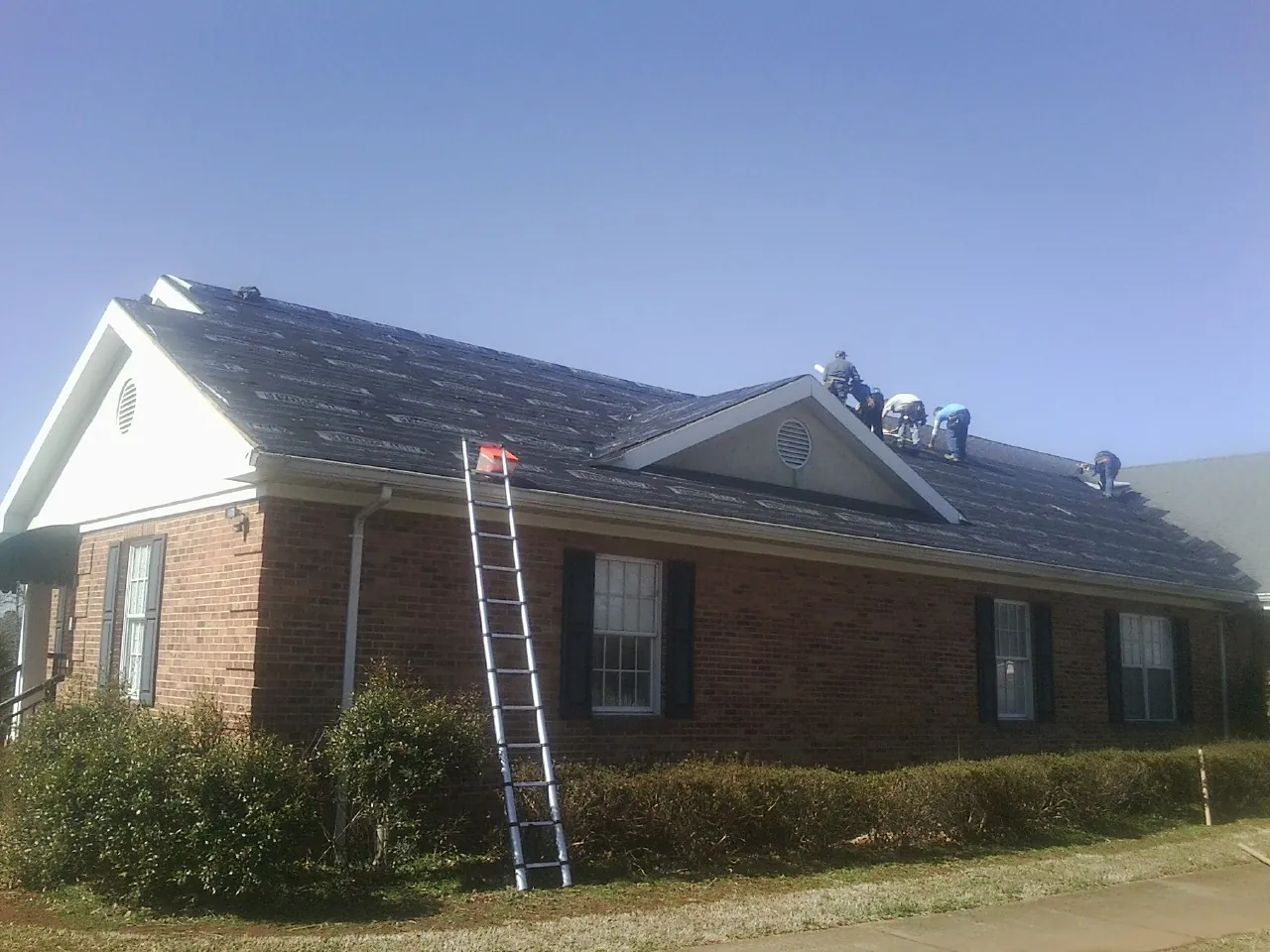Crew tearing shingles off a brick ranch roof with a ladder set — Metal Roof Repair work by our crew in Milford Center