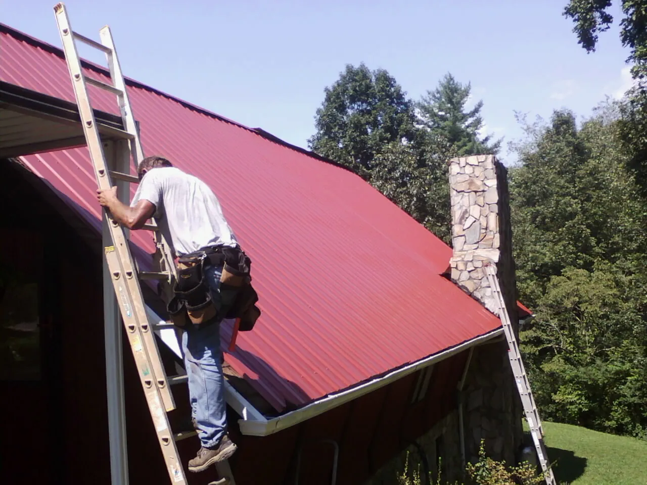 Roofer on a ladder beside a red metal roof and stone chimney — Soffit & Fascia Repair by Roofing Brothers in Milford Center